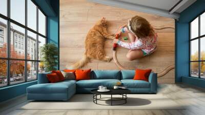 Portrait of cute happy caucasian kid girl and red kitten. Little positive kid sitting and playing on the floor with wooden train road. Warm floor for child and domestic animal. Wall mural