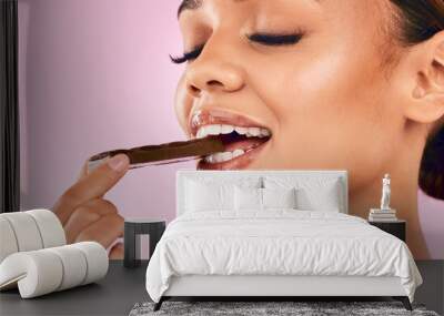 Happy woman, face and eating sweet chocolate, cocoa or delicious dessert against a pink studio background. Closeup of calm female mouth, lips or teeth biting and enjoying tasty sugar food or candy Wall mural