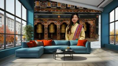 Bhutanese young woman facing the camera with a smile is wearing a kira (national dress) and standing in front of prayer wheels in monastery Wall mural