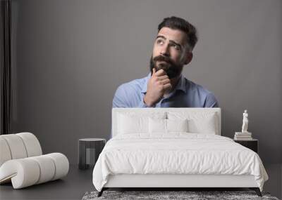 Young adult hipster business man thinking and looking up at copyspace while touching beard against gray studio background.  Wall mural