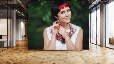 A beautiful girl on a green natural background adjusts her earring with her hand.The Ukrainian bride is dressed in ethnic style.Handmade jewelry.Original set of earrings and a necklace for the holiday Wall mural