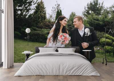 A fashionable groom in a black suit and a beautiful brunette bride in a white dress with a bouquet in her hands are walking in the park holding hands. Wedding portrait of smiling newlyweds. Wall mural