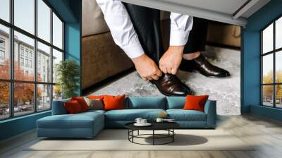 A man, the groom puts leather stylish shiny brown shoes on his feet in socks in the morning, sitting on the sofa and tying his shoelaces with his hands. Closeup photo of a businessman, business. Wall mural