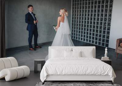 A stylish groom stands near a gray wall and looks at his beautiful bride in a long dress. Delicate and smiling newlyweds in a gray studio. Film effect. Wall mural