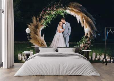 A young groom in a gray suit and a cute, smiling blonde bride embrace, standing on the green grass, near a round wooden arch decorated with reeds, flowers, candles. Night wedding portrait. Wall mural