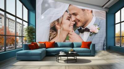 A young groom in a gray suit and a cute, smiling blonde bride with a bouquet are hugging tenderly under a long veil. Wedding portrait of newlyweds in love. Nice photo. Wall mural