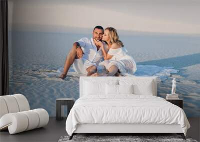 The bearded man and the blonde girl are hugging while sitting on the white sand and enjoying the sunset. Desert and newlyweds. Love story of cheerful and loving people. Vacation, honeymoon. Wall mural