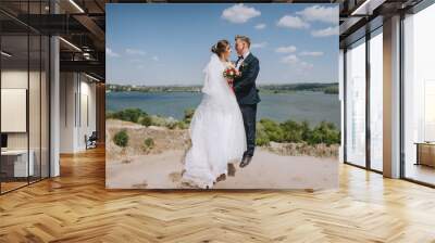 Wedding portrait of newlyweds in love in nature. Stylish groom in a blue suit and a cute bride in a long white dress embrace against the backdrop of rocks, cliffs, sky and river. Wall mural