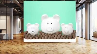 Three piggy banks in a row one large two small behind a wall of coins stacked in piles. Wall of savings. Dollar signs in their eyes. Light green background with copy space. Wall mural