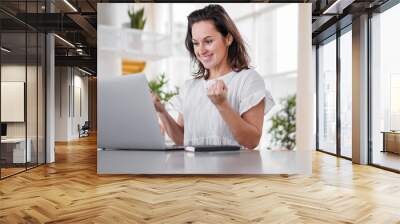 happy joyful successful remote working woman cheerful with fists infront of laptop or notebook in casual outfit sitting on work desk in modern loft living room home office Wall mural