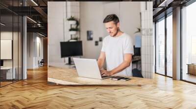 happy smiling remote online working man typing and with laptop, mobile phone and notebook in white shirt standing up in front of a work desk in an coworking office Wall mural