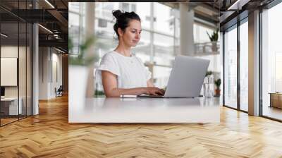 happy smiling remote working dark haired woman typing on laptop or notebook in casual outfit sitting on her work desk in her modern bright loft living room home office with many windows Wall mural