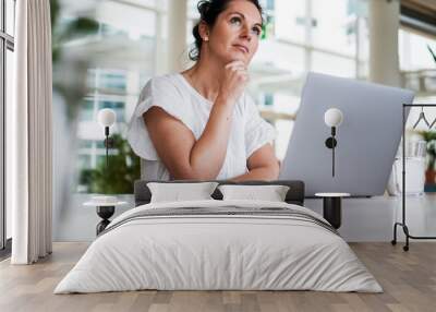 thoughtful brooding remote working dark haired woman sitting infront of a laptop or notebook in casual outfit on her work desk in her modern airy bright living room home office with many windows
 Wall mural