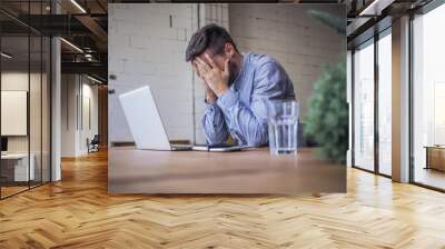 worried and disappointed remote online working man in casual outfit with laptop sitting in an coworking / home office at a work desk Wall mural