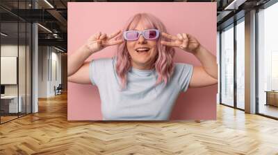 Smiling woman posing in glasses makes v sign shows peace gesture isolated over pink background. Wall mural