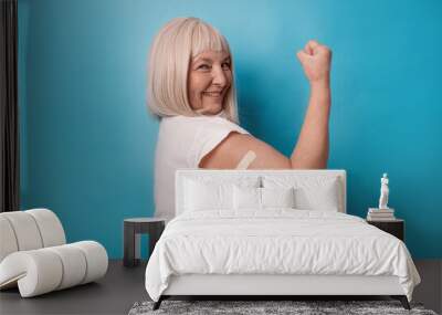 Vaccinated 50s mature older senior woman getting a vaccine protection the coronavirus. Happy female showing arm with bandage after receiving vaccination over blue background. 2024 Wall mural