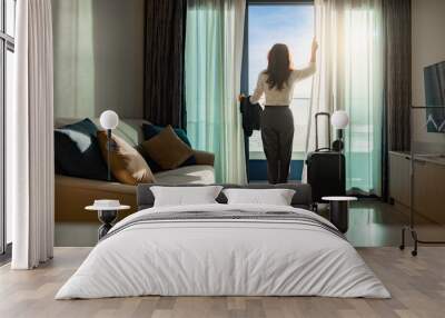 Confident businesswoman wearing white shirt and holding suit with suitcase stands in a hotel room while looking at the view from window Beautiful sunset. Woman enjoying her holiday in a luxury hotel. Wall mural