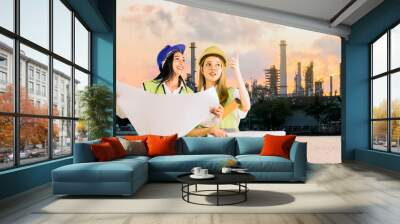 Engineers at work. Two Beautiful asian businesswoman engineer holding the blueprint and wearing the safety helmet on background blurred oil refinery at sunset. Wall mural