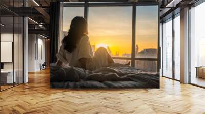 The sun shines through window in the morning. Beautiful young woman sitting on bed. Happy young girl greets new day with warm sunlight flare. Selective focus Wall mural