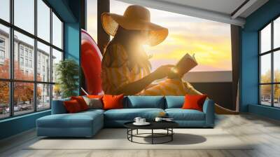 Woman in hat reading interesting book, travel guide books, novel with Rubber swim tube of heart shape while relaxing on floor at home. Sun shines on her from the big window with warm sunlight flare.  Wall mural