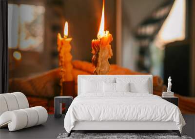 Close-up of two lit candles with melted wax, creating a cozy ambiance against a blurred background of a softly lit room at sunset, capturing the warmth and tranquility of a quiet evening Wall mural