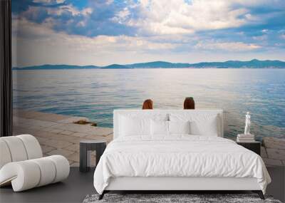 Two young girls with backpacks sitting on the stone near the sea shore, their hands leaning against stone pavement, looking at the hill range in the distance. Town of Zadar, Croatia Wall mural