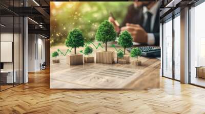 A businessman working at his desk with a calculator and a pen, with small trees growing on wooden blocks in front of him, representing growth and sustainability. Wall mural