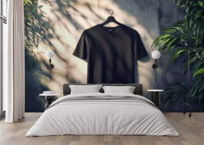 Blank black t-shirt hanging on a hanger against a concrete wall with green plants and sunlight shadows. Wall mural