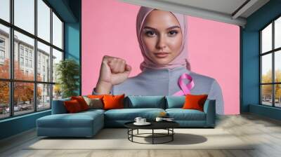 Bald pretty arab woman showing fist, pink ribbon lapel pin - symbol of the fight against breast cancer on her clothes, bright solid background Wall mural