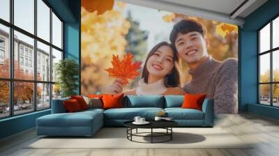 Handsome lovely asian woman and man couple in the fall season autumn landscape sunny day, trees and leaves background, holding colorful maple leafs Wall mural