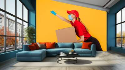 agitated young delivery woman, in a red uniform, wearing a medical protective mask and gloves, running and holding a cardboard box isolated on a yellow background. Express delivery, delivery service Wall mural