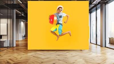 Full-length portrait of a happy excited teenager in sunglasses and a summer hat, having fun and jumping, holding an inflatable sea ball on a yellow background Wall mural