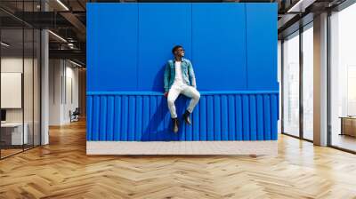 Handsome african male fashion model in sunglasses smiling and posing while standing near blue wall outdoors Wall mural