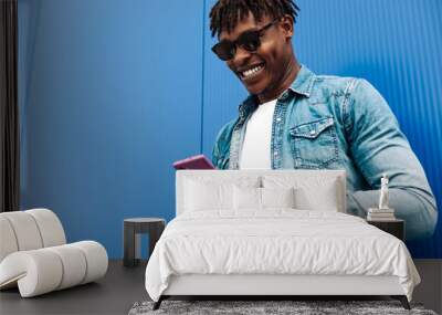 happy curly african american man wearing sunglasses using mobile phone and smiling against blue wall background Wall mural