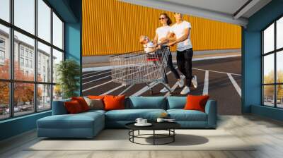 Happy family, man and woman having fun while rolling their baby in shopping trolley, outdoors, family shopping concept Wall mural