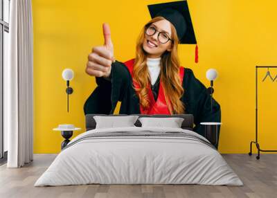 Happy graduate in a graduation cap, gives a thumbs up and smiles on an isolated yellow background Wall mural