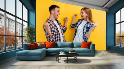 Shocked happy young man and woman, in checkered shirts and white t-shirts, celebrate the victory, showing a victory gesture, on a yellow background Wall mural