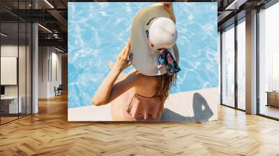 The girl holds a light hat, covering her whole body with her hands, sitting by the pool in the sun with her back Wall mural