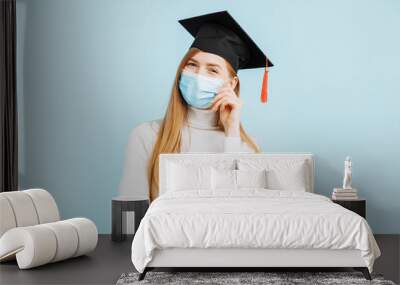 thinking girl graduate in a medical mask, student in a graduation hat thinks and looks to the side on a blue background Wall mural