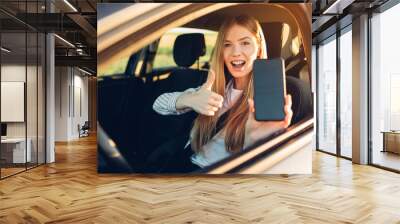 Young happy woman driving a car, she shows a blank phone screen and a thumb up Wall mural