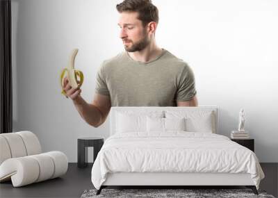 Handsome Young Man in Green Shirt Holding a Banana Symbolizing Male Genitals, Prostate, and Sexuality, Looking at It with a Skeptical Expression Against White Background Wall mural