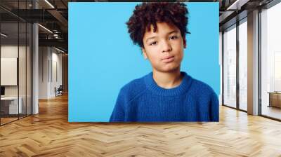 A joyful young african american boy with curly hair wearing a blue sweater against a vibrant blue background Wall mural