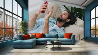 A man with a beard lies on the couch during the day at home and looks at his phone relaxing on his day off, a man gambling on the stock market online on his phone Wall mural