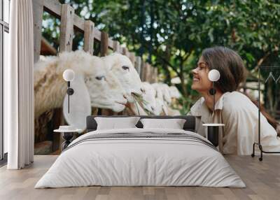 A woman petting a sheep in a pen with a fence in the background and trees in the background Wall mural