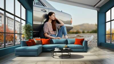 A young woman sits on the ground near her car on the side of the road and looks at the sunset Wall mural