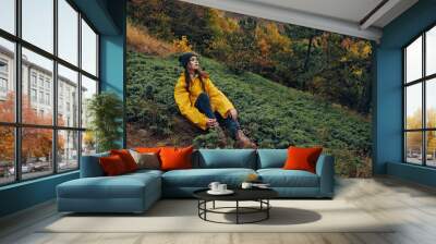 Beautiful young woman in yellow jacket sitting alone on grassy hill surrounded by autumn forest serene nature getaway fashion photo concept Wall mural