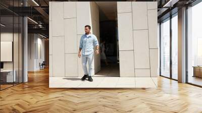 Confident young man in casual attire walking out of modern building, embracing a sunny day Wall mural