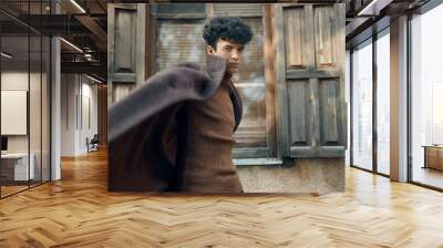 Fashion young man in brown sweater standing in front of old building with open and wooden shuttered window Wall mural