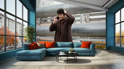 Fashionable man in a brown coat talking on a cell phone on a bridge with a large metal pipe in the background Wall mural