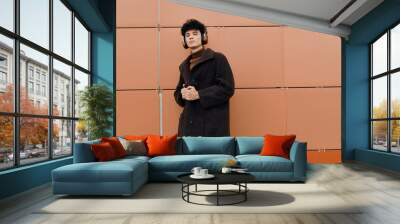 Fashionable young man in a coat and scarf listening to music with headphones against an orange wall Wall mural